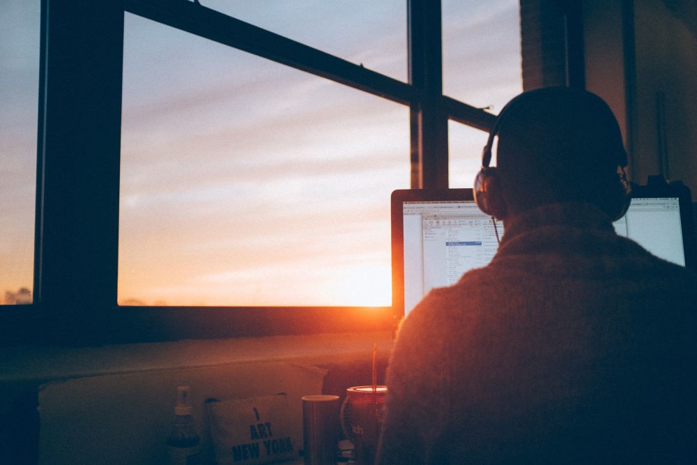 Mężczyzna siedzi w słuchawkach przed komputerem. Za oknem zachód słońca.